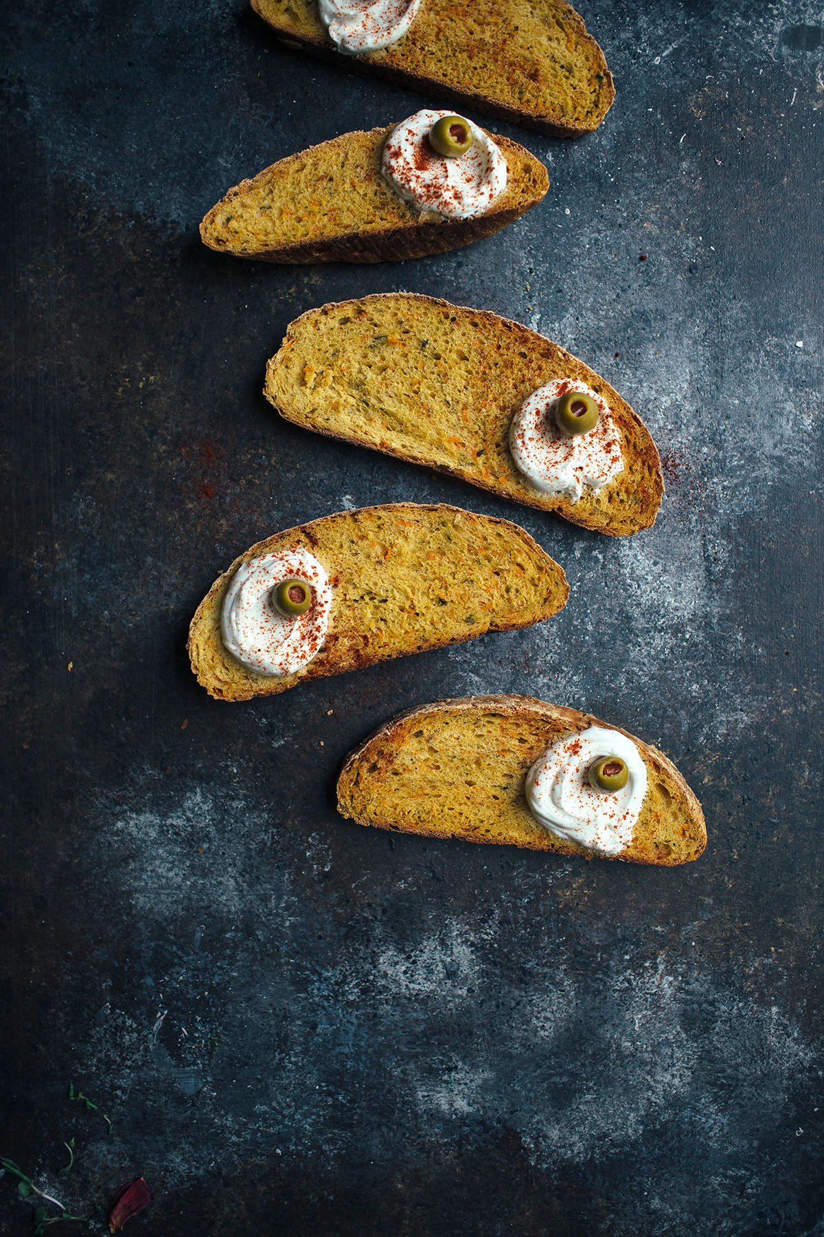 five slices of vegetable bread topped with yogurt and olives on dark background