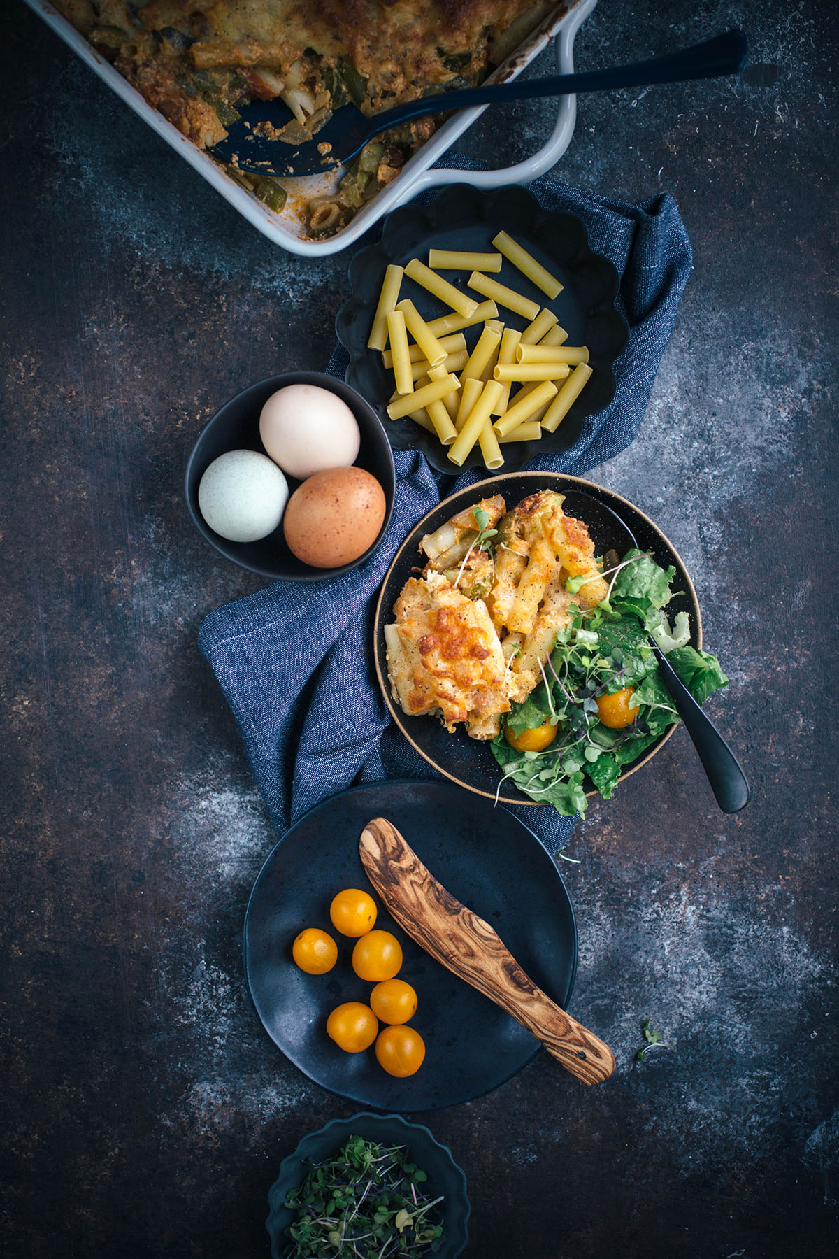 Vegetarian Ziti with side salad on dark plate next to dried pasta, eggs, and tomaotes