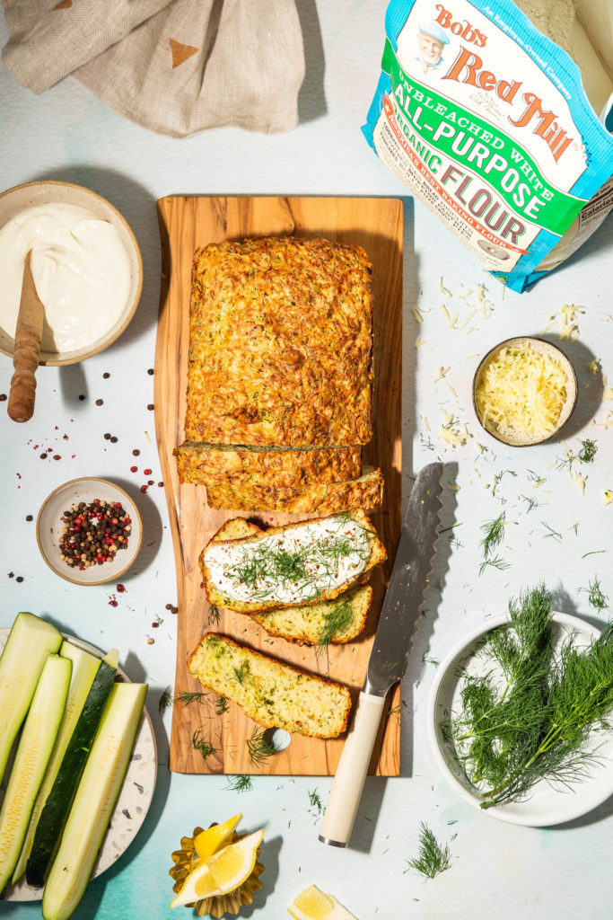 partially sliced loaf of Zucchini cheese Bread on wood cutting board surrounded by ingredients 