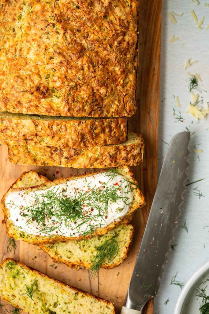 slices of zucchini cheesy bread on wood cutting board topped with whipped goat cheese and dill