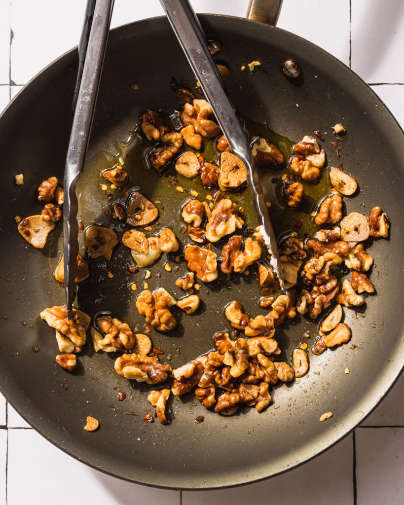 walnuts, oil, and red pepper flakes in skillet with tongs